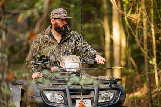 A bearded man in a Non-Typical Endurance Hooded Full Zip Jacket with Scent Control by Nontypical rides an ATV through a sunlit forest, with an arrow attached to the front.
