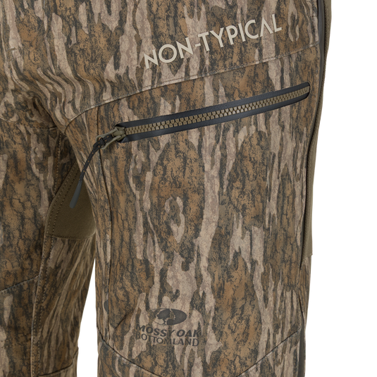 Close-up of Non-Typical Pursuit Softshell Pant with Agion Active X2 windproof hunting pants in camouflage, featuring a zippered thigh pocket, the text NON-TYPICAL, and a MOSSY OAK BOTTOMLAND logo.