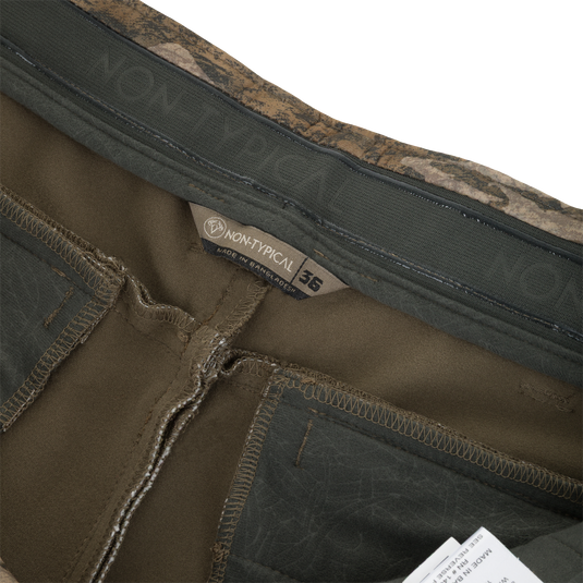 Close-up of the interior waistband of these olive green Nontypical Non-Typical Pursuit Softshell Pant with Agion Active X2 pants, featuring a camouflage-patterned trim, a size 36 label, and a brand tag.