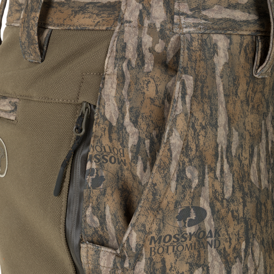 Close-up of olive green and camouflage Non-Typical Pursuit Softshell Pant with Agion Active X2 hunting pants featuring a side zipper pocket and printed Mossy Oak Bottomland branding.
