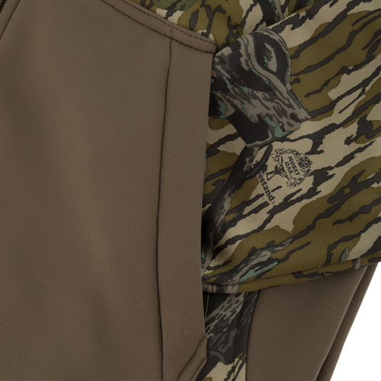 Close-up of a zippered pocket on a Nontypical Youth Endurance Full Zip Jacket with Hood, featuring a solid olive panel and a camouflage pattern with a Mossy Oak logo.
