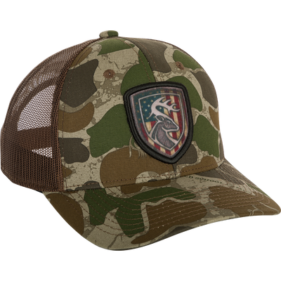 A camouflage hat with an Americana shield patch on it, made of 100% cotton twill front panels and breathable mesh on the back. Features a snap closure and the Non-Typical logo on the front.