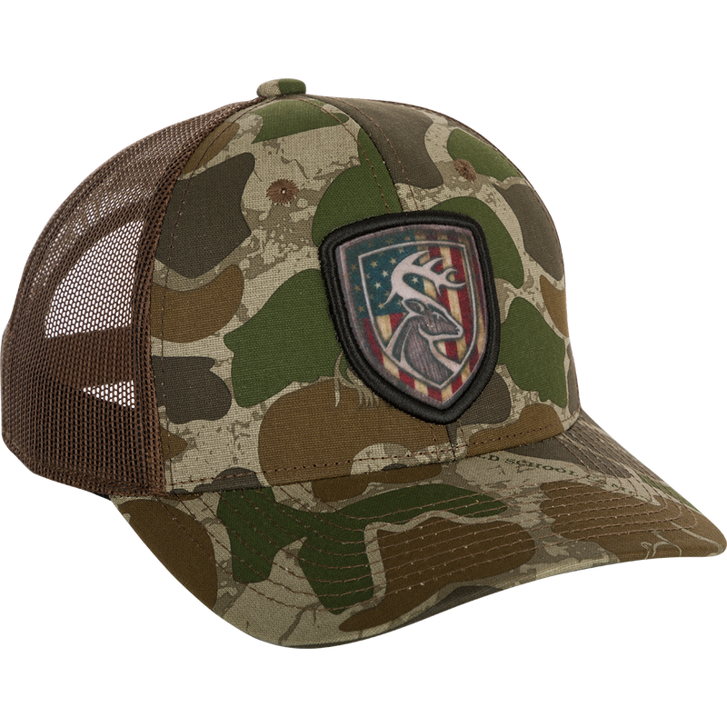 A camouflage hat with an Americana shield patch on it, made of 100% cotton twill front panels and breathable mesh on the back. Features a snap closure and the Non-Typical logo on the front.