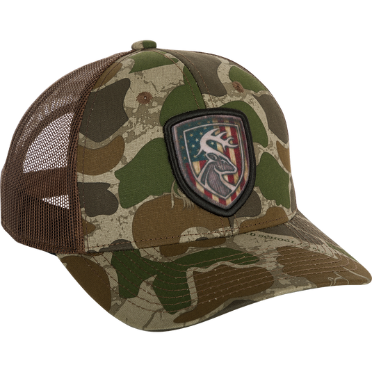 A camouflage hat with an Americana shield patch on it, made of 100% cotton twill front panels and breathable mesh on the back. Features a snap closure and the Non-Typical logo on the front.
