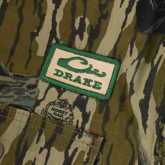 The Campfire Canvas Bomber Jacket by Lifestyle features rugged green and brown camo, a rectangular Drake patch, and a small logo above the Mossy Oak-labeled pocket.
