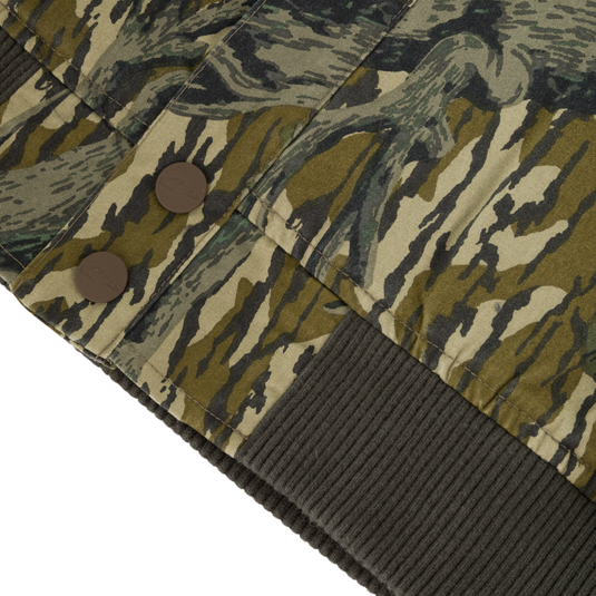 Close-up of the Campfire Canvas Bomber Jacket by Lifestyle in a camo pattern, with two brown buttons, a ribbed dark green hem, and fabric featuring green, brown, and beige abstract shapes.