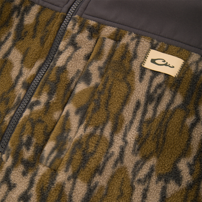 Close-up of a Camp Fleece Over Under Quarter Zip in a brown and green camouflage pattern with a solid dark-colored chest panel and a small brand logo patch.