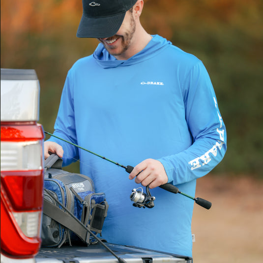 A man in a blue Performance Hoodie Solid, holding a fishing pole. Fabric: Cooling, UPF 50, Moisture Wicking, Breathable Stretch, Quick Drying. From Drake Waterfowl's versatile hunting and outdoor apparel line.
