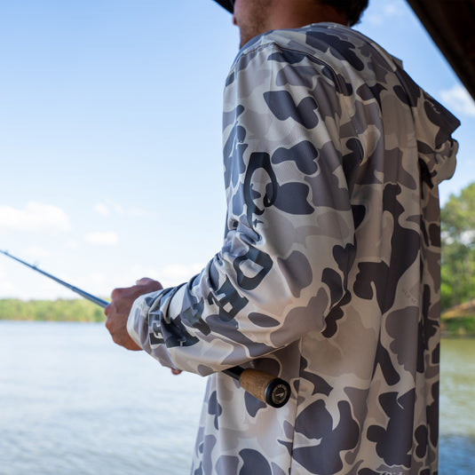 A man fishing on a lake, holding a fishing pole. The Drake Performance Hoodie: Cooling, UPF 50, Moisture Wicking, Breathable, Quick Drying. Versatile for all weather conditions. Materials: 92% Polyester/8% Spandex Jersey.