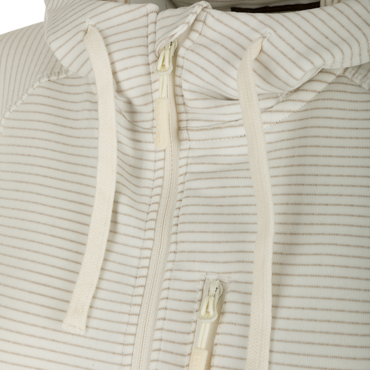A close up of the Women's MST Breathlite Hoodie, featuring a white and tan striped fabric. The hoodie has a zipper and a vertical YKK zippered left chest pocket. It also has long sleeves with thumb loops and a hood with adjustable drawstring closure. The hoodie is made with a midweight 94% Polyester Fleece and 6% Spandex material, providing comfort and durability. Perfect for outdoor activities.
