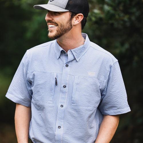 A man in a Classic Seersucker Stripe Button-Down Short Sleeve Shirt with button-down collar, chest pockets, and vented cape back, embodying Drake Waterfowl's high-quality hunting and casual apparel.