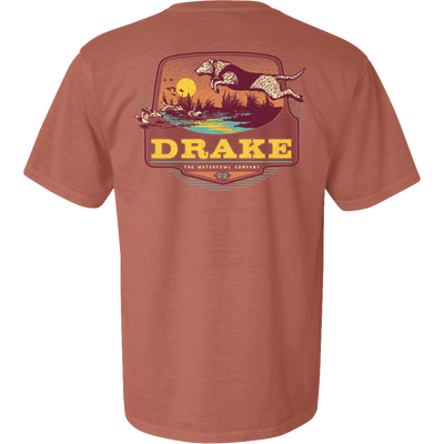 Don’t Let Em Behind You T-Shirt featuring a hunting dog graphic leaping over a marsh with ducks, sunset backdrop, and Drake logo on the back.