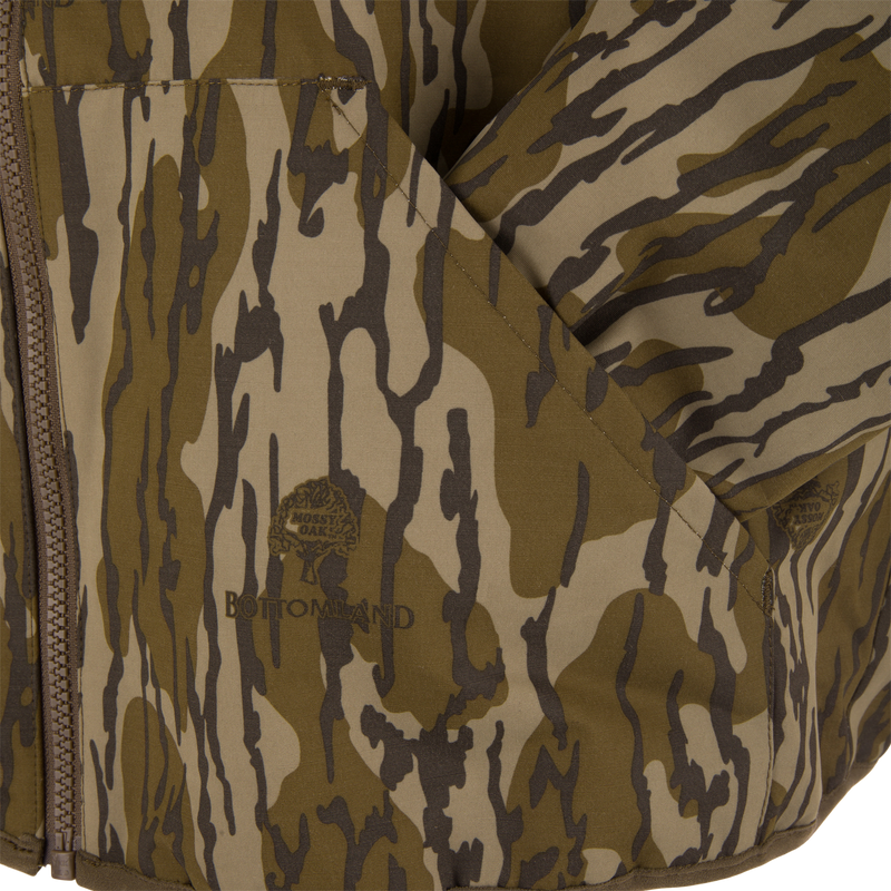 Close-up of the Throwback 80 Sport 3-in-1 Jacket in Mossy Oak Bottomland pattern, showcasing its zip closure, fold-over pocket, and detailed logo.