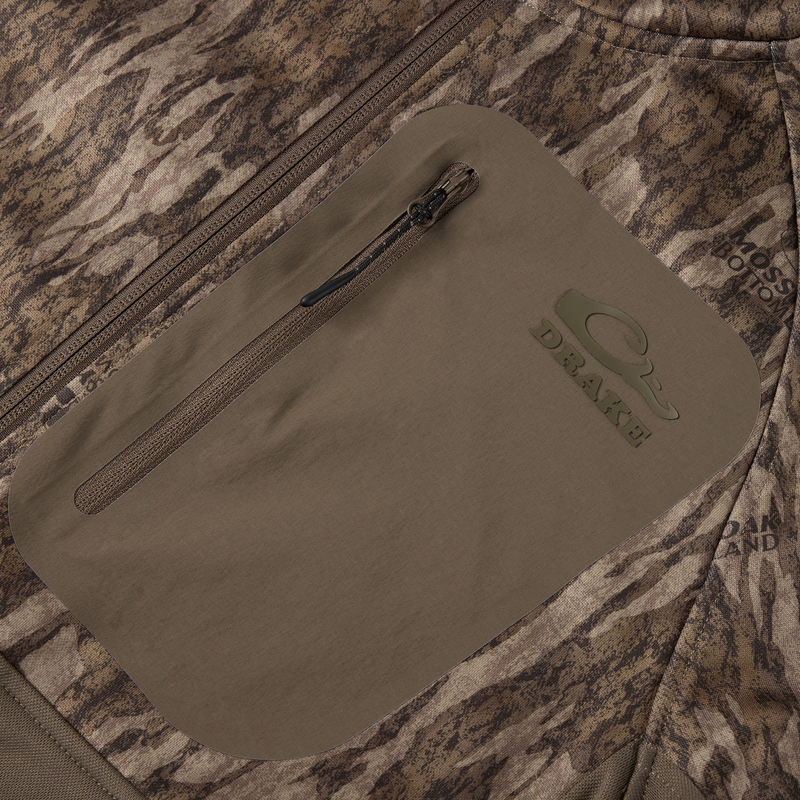 Close-up of MST Offset 2.0 Half Zip Hoodie's zippered, water-resistant chest pocket with Drake logo and Mossy Oak Bottomland camo, showcasing rugged outdoor design.