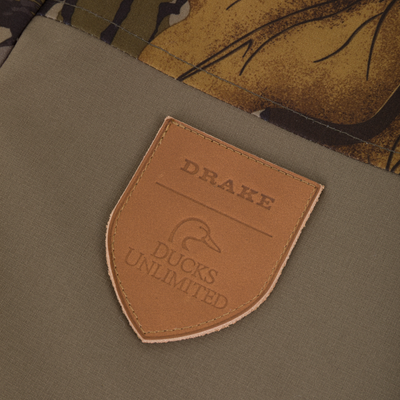 A brown leather shield-shaped patch on a camouflage and olive green Drake X Ducks Unlimited LST BMF Jacket with Primaloft, embossed with the text Drake Ducks Unlimited and a duck silhouette.