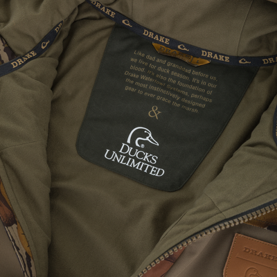 Interior view of a Drake X Ducks Unlimited LST BMF Jacket with Primaloft showing the neck label printed with a Ducks Unlimited logo and descriptive text about waterfowl conservation heritage.