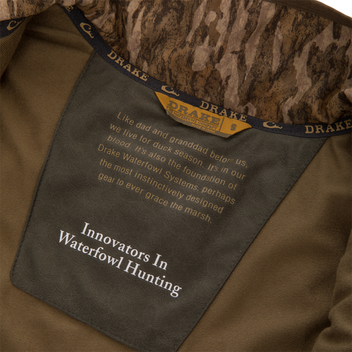 Inside view of Drake Waterfowl LST BMF Vest with Primaloft, showcasing camo collar, size tag, and product slogan emphasizing innovation and warmth for hunting.