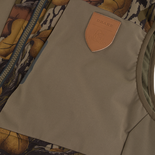 Close-up of a Drake X Ducks Unlimited LST BMF Vest with Primaloft, showing a tan chest panel with a leather-style shield patch embossed with a duck logo and the words Drake Ducks Unlimited.