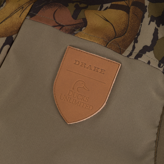 A tan, shield-shaped leather patch on a Drake X Ducks Unlimited LST BMF Vest with Primaloft featuring the embossed text Drake and Ducks Unlimited above a duck head silhouette.
