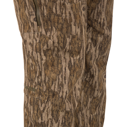 Close-up of MST CommandXT Softshell Pant in wood-grain camo, featuring a zippered side pocket, OAKLAND branding, and DRAKE logo, highlighting mobility and protection.