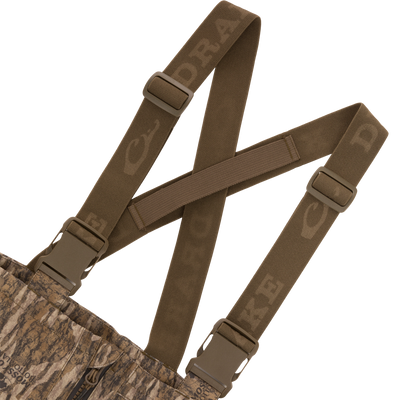 LST CommandXT Bib with Primaloft shows camo pattern, X-crossed brown suspenders, side-release buckles, and Mossy Oak Bottomland logo near zipper, ideal for extreme hunting conditions.