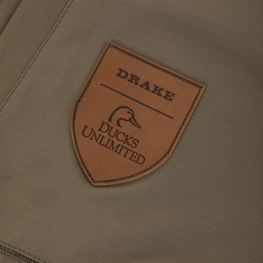 Brown shield-shaped leather patch featuring the Drake logo and Ducks Unlimited text, stitched onto the olive green fabric of a Drake X Ducks Unlimited LST BMF Pit Bib with Primaloft.