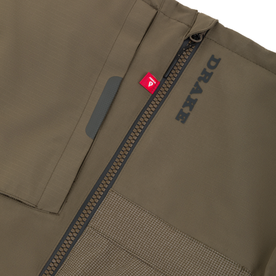 Close-up of an olive green Drake X Ducks Unlimited LST BMF Pit Bib with Primaloft, featuring a vertical zipper, a small red Primaloft tag, and black text branding.