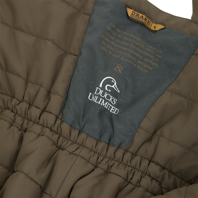 Olive green quilted Drake X Ducks Unlimited LST BMF Pit Bib with Primaloft, featuring a central grey patch with the Ducks Unlimited logo and an inspirational quote about waterfowl hunting.