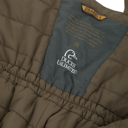 Olive green quilted Drake X Ducks Unlimited LST BMF Pit Bib with Primaloft, featuring a central grey patch with the Ducks Unlimited logo and an inspirational quote about waterfowl hunting.