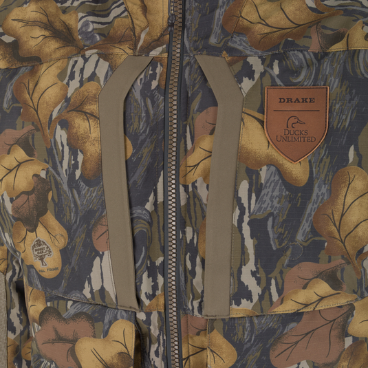 Close-up of a Drake X Ducks Unlimited LST BMF 3-in-1 Pit Jacket with Primaloft camouflage jacket with a front zipper, tan trim, and a leather patch featuring a duck logo.