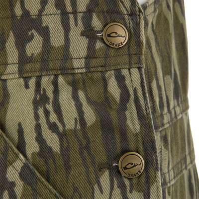 Close-up of Throwback 80 Field Bib's camo fabric with detailed stitching and a brass button embossed with DRAKE, showcasing durability and classic workwear style.