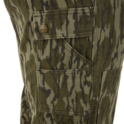 Close-up of a Throwback80 Field Bib's flap pocket with a brass snap button, showcasing durable camo print fabric and Drake branding for rugged field use.