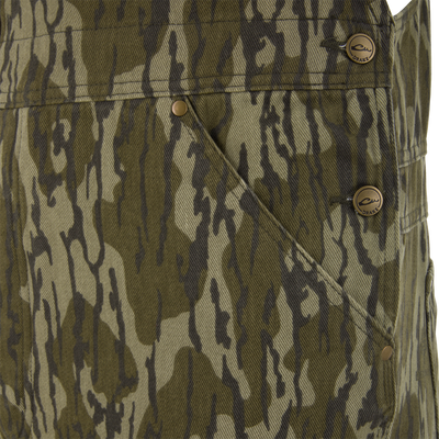 Close-up of Throwback 80 Field Bib's camouflage fabric, showcasing diagonal pocket with metal snap button stamped DRAKE, highlighting durable design and workwear inspiration.