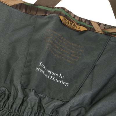 Drake Waterfowl BMF Uninsulated Pit Bib's interior features Innovators In Waterfowl Hunting slogan, adjustable camo-trimmed straps, and elastic waist, highlighting its durable, high-performance design.