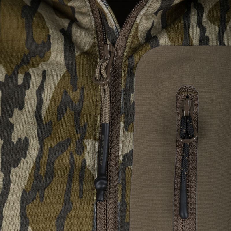 Close-up of Drake Waterfowl's Grid Fleece Half Zip Hoodie, highlighting camo fabric, main zipper with corded pull, and water-resistant chest pocket with branded pull.