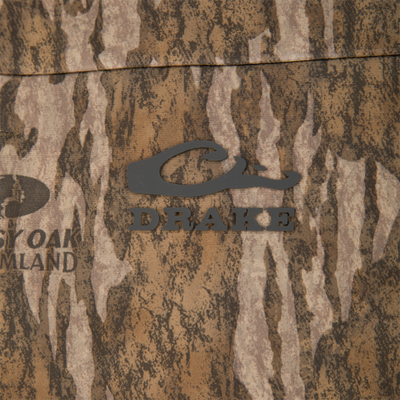 Close-up of the MST CommandXT Softshell Vest featuring a camouflage pattern and DRAKE logo, highlighting its water-resistant, windproof, and flexible design for outdoor activities.