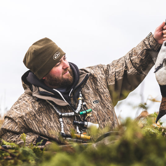 A hunter in camouflage holds a duck, showcasing the LST Refuge™ 3.0 3-in-1 Jacket's versatility and protection. Features include waterproof fabric, Eqwader technology, and multiple pockets for hunting essentials.