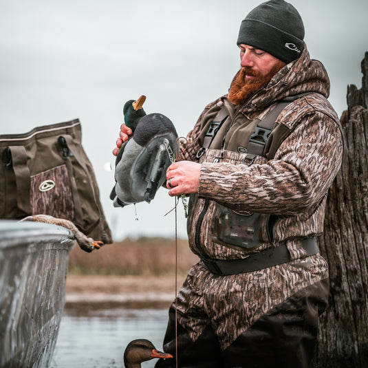 A man in a camouflage hoodie holding ducks in water, showcasing the MST Endurance Soft Shell Hoodie by Drake Waterfowl. Features Magnattach™ pocket, zipped lower pockets, mesh-lined sleeves, and fleece-lined hood.