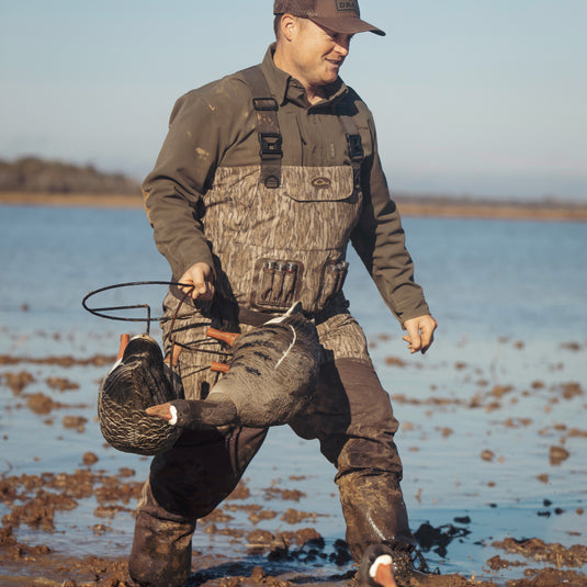 A man in hunting gear carrying decoys in mud, showcasing the Insulated Guardian Elite Vanguard Breathable Waders by Drake Waterfowl. Waterproof, windproof, with LokDown insulation and Thinsulate boots for warmth and comfort.