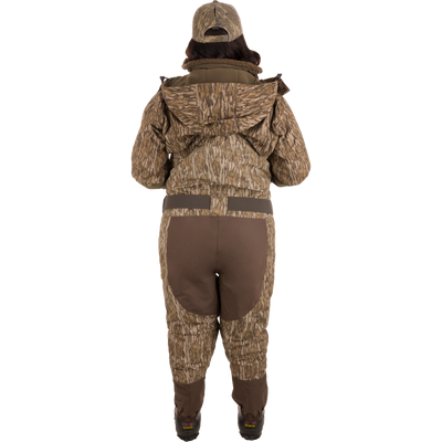A person wearing Women’s Insulated Guardian Elite Vanguard Breathable Waders with a camouflage pattern, featuring reinforced seams, handwarmer pockets, and Thinsulate Buckshot Mud boots.