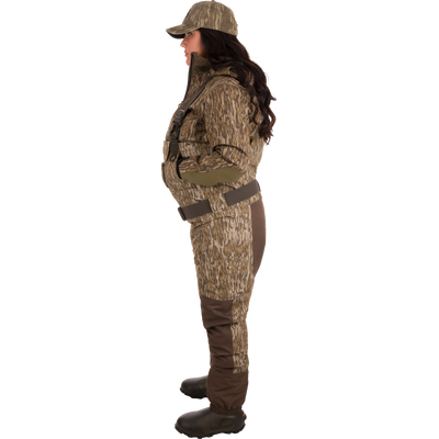 Woman wearing Women’s Insulated Guardian Elite Vanguard Breathable Waders with camouflage outfit, featuring reinforced seams, handwarmer pockets, front cargo pouch, and Thinsulate Buckshot Mud boots.