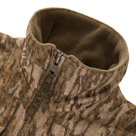 Close-up of the MST Youth Eqwader Quarter Zip Jacket, featuring a camouflaged, fleece-lined design with a tall collar, branded DRAKE zipper pull, and patented waterproof technology.