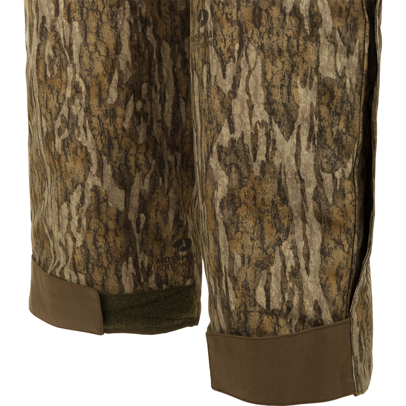 A pair of camouflage Guardian Elite Bibs, perfect for hunters seeking warmth in frigid weather conditions. Waterproof, windproof, and breathable, with reinforced knees and seat. Features include chest pocket, handwarmer pockets, and full-length side leg zippers.