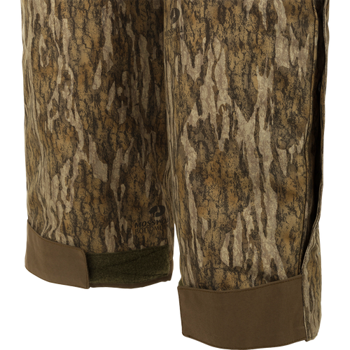 A pair of camouflage Guardian Elite Bibs, perfect for hunters seeking warmth in frigid weather conditions. Waterproof, windproof, and breathable, with reinforced knees and seat. Features include chest pocket, handwarmer pockets, and full-length side leg zippers.