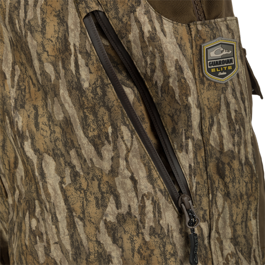 A close-up of the LST Guardian Elite Bibs, featuring a jacket with a zipper, patch, and brown knitted fabric. Perfect for hunters seeking warmth in frigid weather conditions. Waterproof, windproof, and breathable. Full-length side leg zippers for easy on/off. Magnattach™ chest pocket, call separator, handwarmer pockets, and more.