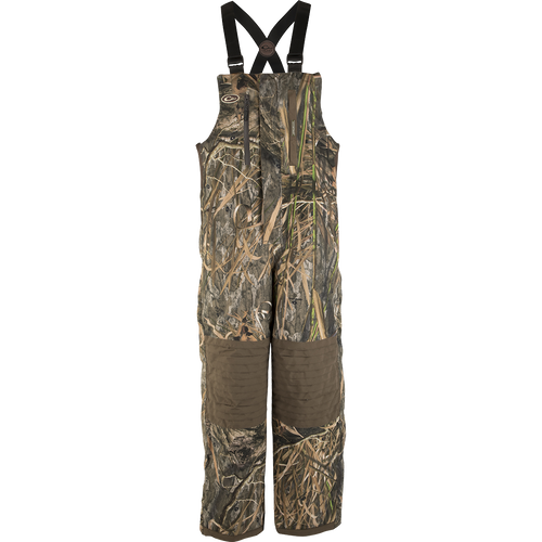 A product image of the EST Guardian Elite™ Bib Shell Weight, featuring a camouflage overall with straps, perfect for hunters seeking protection in all weather conditions. The bib is made of 100% waterproof/windproof/breathable Guardian Elite™ 3-layer pro wader fabric, with reinforced knees, seat, and wear areas. It includes adjustable shoulder straps, Magnattach™ chest pockets, and easy on/off full-length side zippers. Ideal for use in a ground blind.