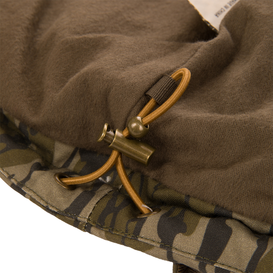 Close-up of the Waterfowl LST Insulated Waxed Cotton Jacket hem featuring an adjustable elastic drawstring cord with a metal toggle fastener against a brown fleece lining and camouflage waxed cotton exterior.