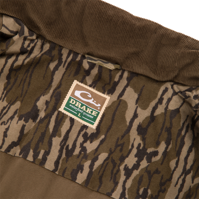 Interior of a Waterfowl LST Insulated Waxed Cotton Jacket featuring a brown corduroy collar and a camo-print lining with a brand label reading Drake Waterfowl Systems and size L.