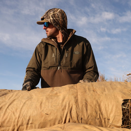 A man in a camouflage wearing sunglasses stands by hay. MST Guardian Eqwader Flex Fleece 1/4 Zip Jacket features waterproof Guardian Flex™ upper, fleece lower, and elbow protection.