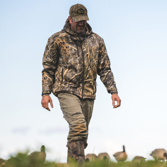 A man in MST Hole Shot Hooded Windproof Eqwader Jacket, camo, walking in a field. Sherpa-lined, windproof upper body, high handwarmer pockets, adjustable waist and hood. High-quality hunting gear from Drake Waterfowl.